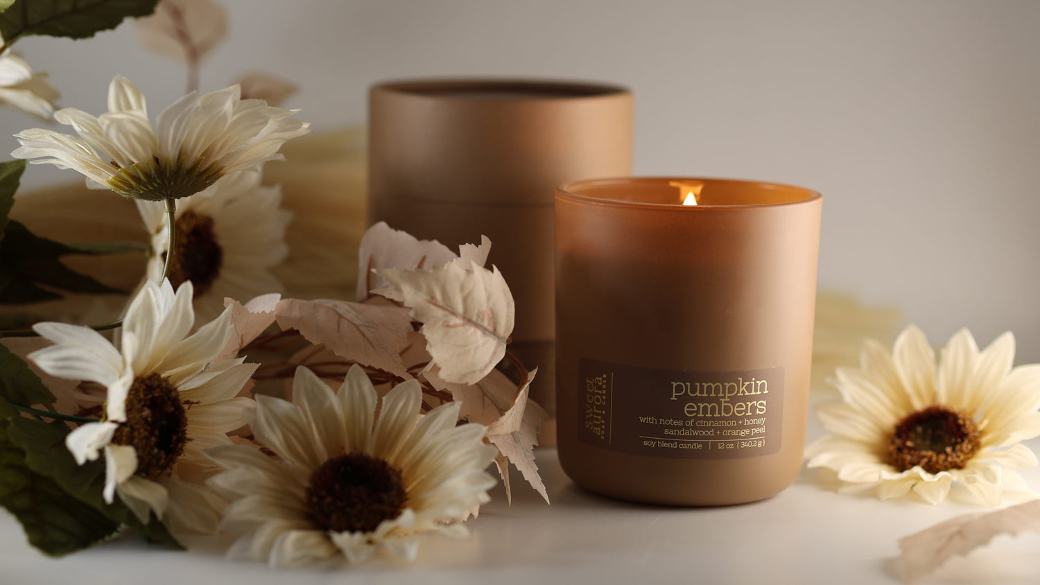 A lit amber-brown candle in a matte glass jar sits on a tabletop with dried flowers and leaves around it. A second unlit candle and a matching cylindrical container are behind it against a soft neutral background.