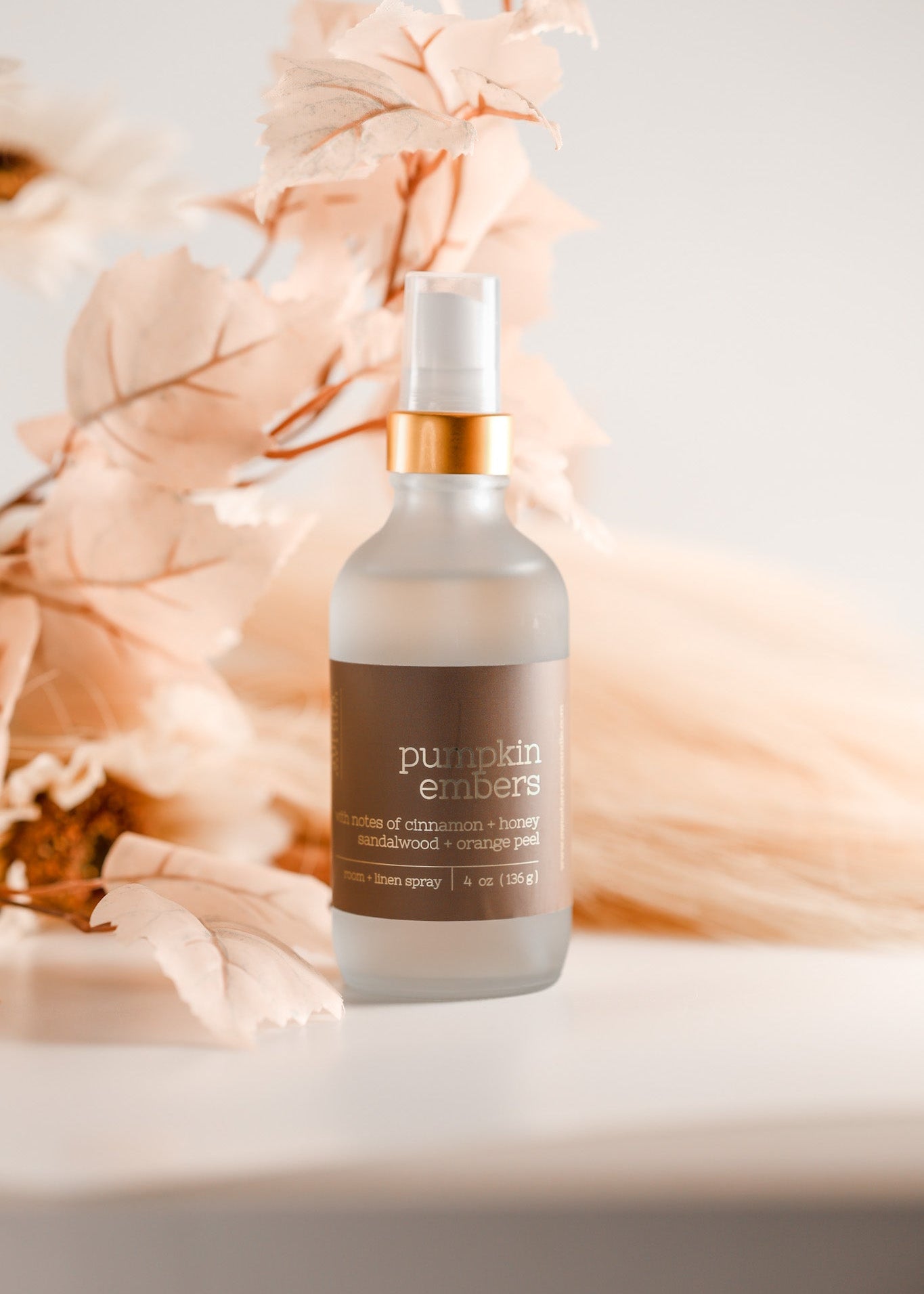 A frosted glass pump bottle labeled 'pumpkin embers' with a white spray top and metallic gold collar, styled with soft peach dried leaves on a light surface against a warm off-white background. Handcrafted by Sweet Aurora Soap & Candle 