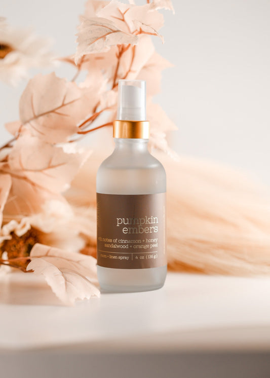 A frosted glass pump bottle labeled 'pumpkin embers' with a white spray top and metallic gold collar, styled with soft peach dried leaves on a light surface against a warm off-white background. Handcrafted by Sweet Aurora Soap & Candle 