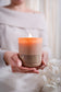A person in a white off-the-shoulder sweater holds a lit candle labeled 'pumpkin embers' in a warm, soft-focus setting.