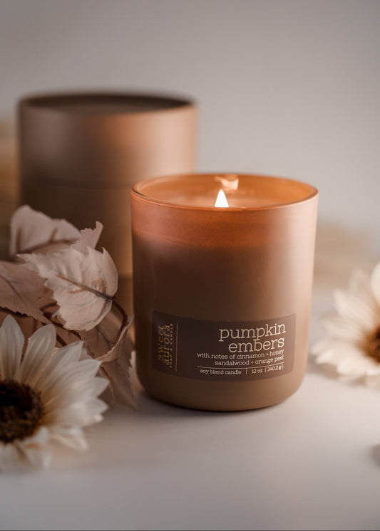A lit amber glass candle jar labeled 'pumpkin embers, by Sweet Aurora Soap & Candle, styled with dried flowers on a light surface with a blurred second jar in the background.