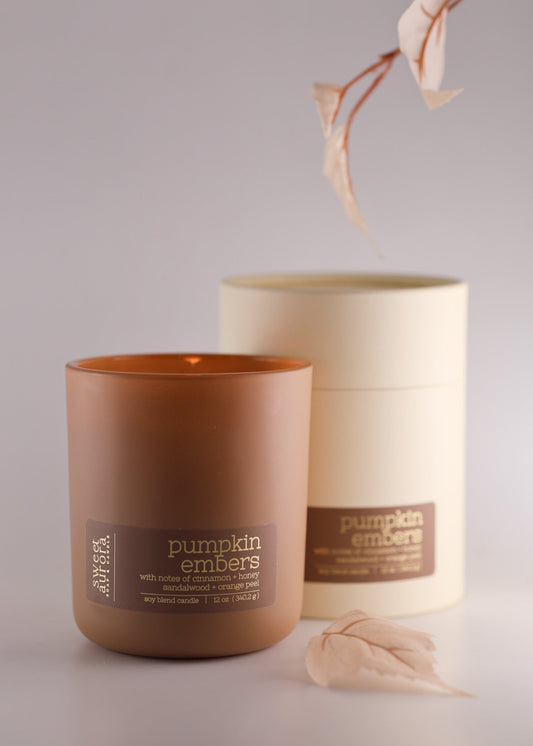 A pumpkin embers scented candle in a frosted amber glass jar with its cream-colored cylindrical packaging box behind it, made by Sweet Aurora Soap and Candle, styled with dried leaves on a light gray background.