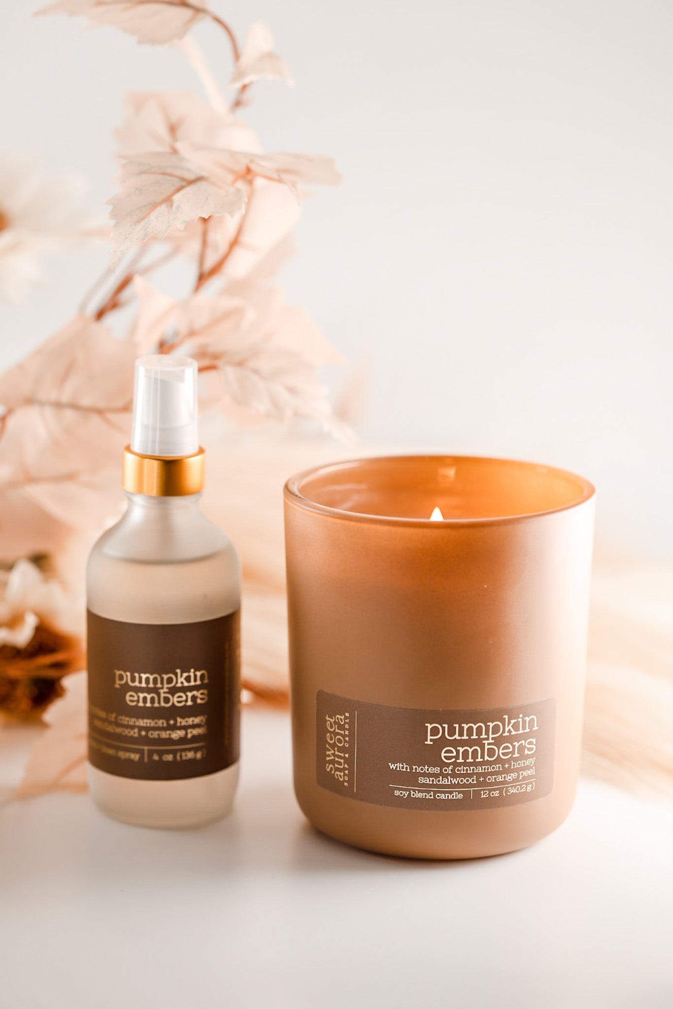 A lit amber glass jar candle and a matching frosted spray bottle labeled 'pumpkin embers' with a gold collar, styled on a white surface with soft dried floral decor in the background.