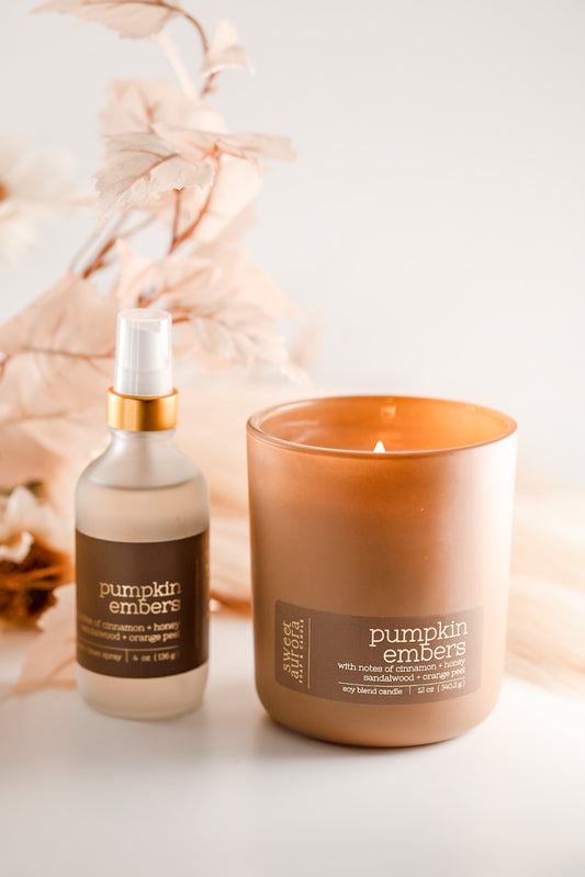 A lit amber glass jar candle and a matching frosted spray bottle labeled 'pumpkin embers' with a gold collar, styled on a white surface with soft dried floral decor in the background.