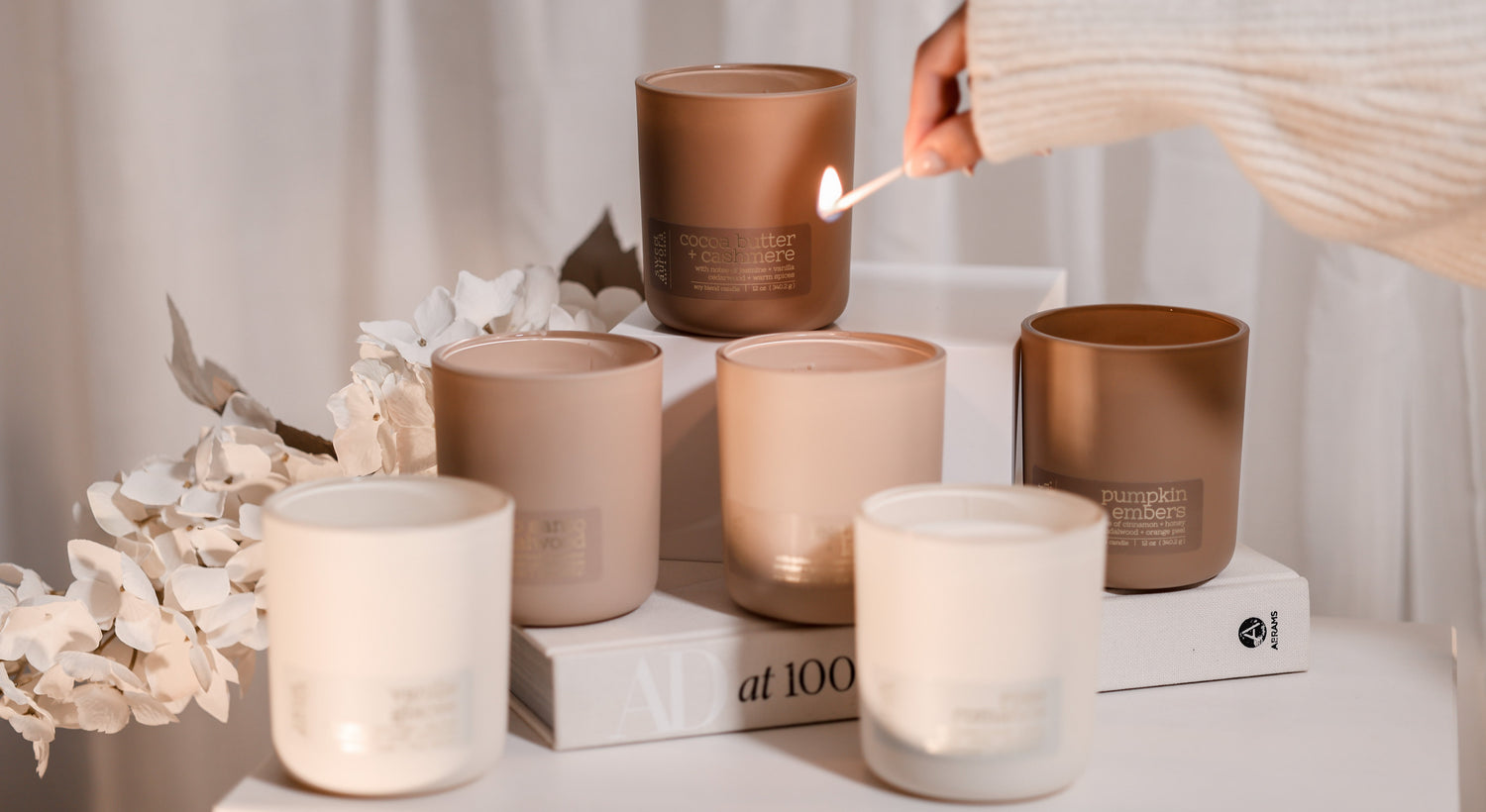 A group of matte candle jars in neutral tones of white, beige, and brown, styled on a white surface with a book and dried flowers. A hand wearing a cream knit sleeve holds a lit match next to one of the brown candles.