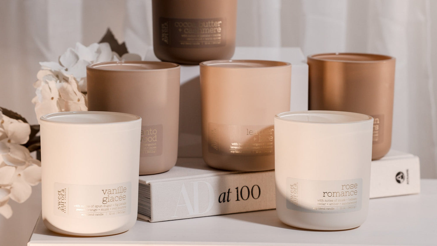 Six matte glass jar candles in neutral earthy tones are arranged on stacked white books with dried flowers. The front candles have white wax and labels reading 'vanilla jasmine' and 'rose romance'; the back candles have warm taupe and brown tones with labels including 'caramel' and 'chocolate'.
